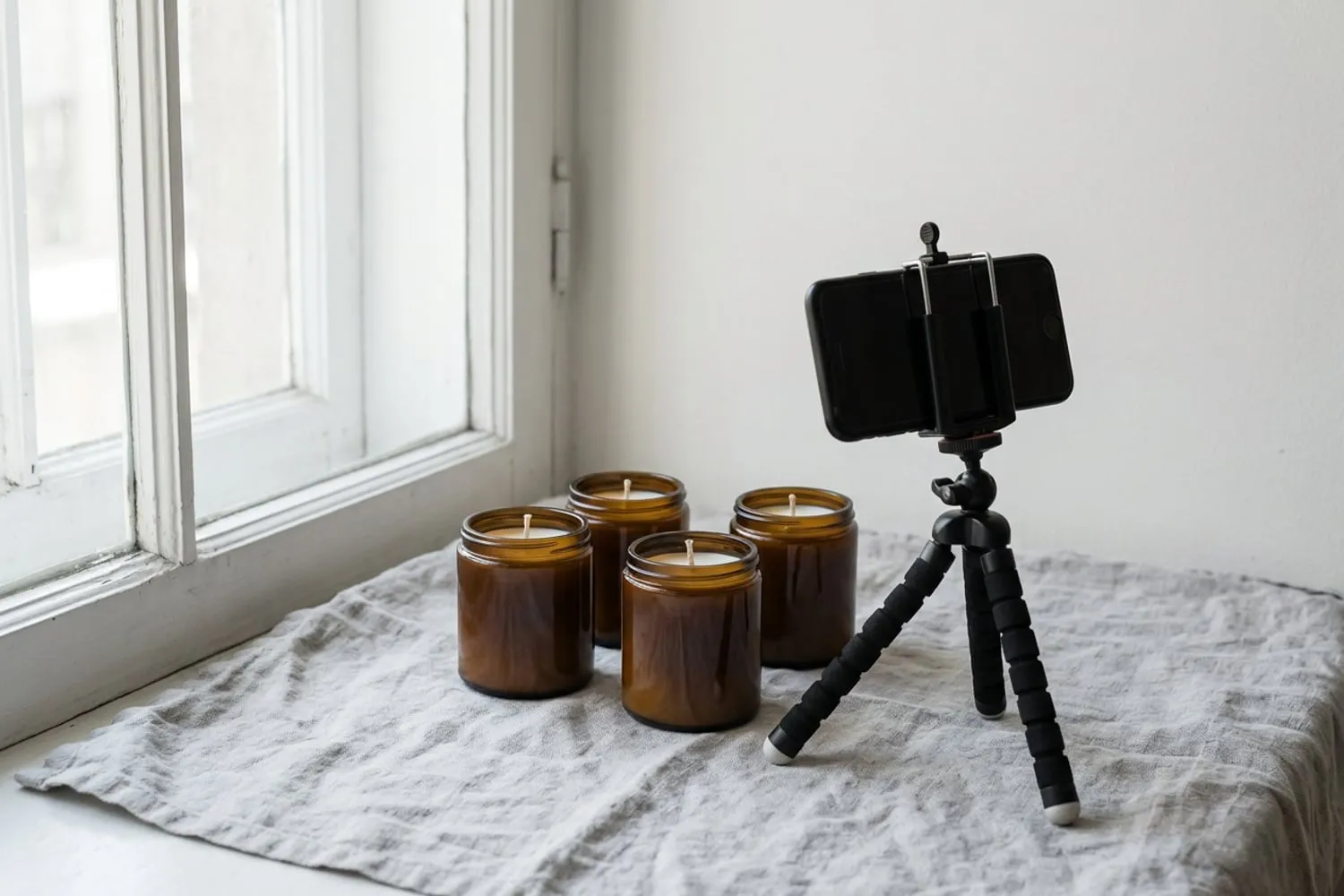 Three handmade candles in plain, unlabeled amber glass jars on a linen cloth by a window, with a phone on a small tripod pointed at them
